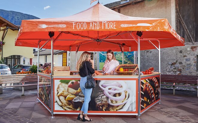 The saleswoman in the hexagonal vegetable stand is offering the customer a carrot to taste. The roof of the market stand is orange and printed with the words "FOOD AND MORE"; the panels, on the other hand, are printed with food all over.