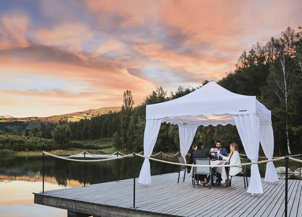 The gazebo kit Royal is located at the footbridge during sunset. Under the gazebo 4 guests are enjoying their dinner. 