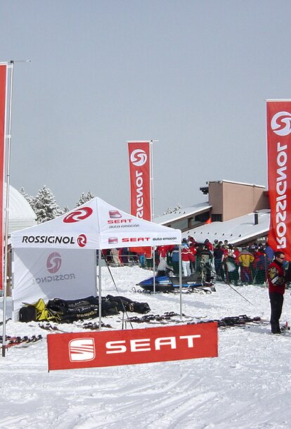 In a ski resort there is a 3x3 m folding gazebo with a white roof and a white sidewall printed in red. There are three large red flags around the gazebo. In the background there are several skiers and buildings.