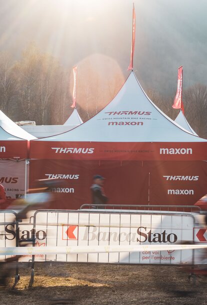 The picture shows three folding gazebos with red roof flags of the Thömus Maxon mountain bike team. The tents have white roofs and red sidewalls. In the background you can see three more roof peaks with the flags.