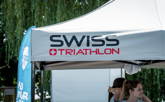 La imagen muestra el techo de una carpa plegable gris de 3x3 m. Lleva impreso el logotipo de Swiss Triathlon.